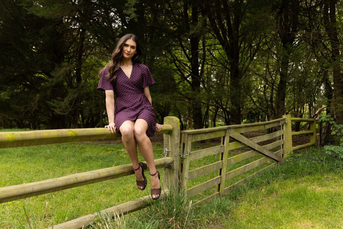 Model in a plum spot wrap dress sitting on a farm fence in front of trees